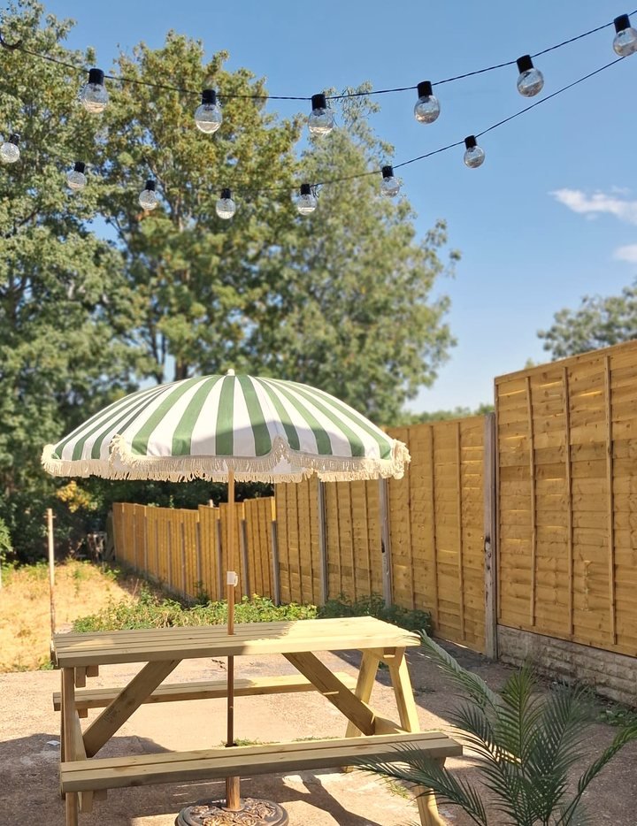 The home&rsquo;s enclosed back garden in summer: a wooden picnic bench under a green and white striped parasol, festoon lights strung overhead, fence panels and trees beyond.