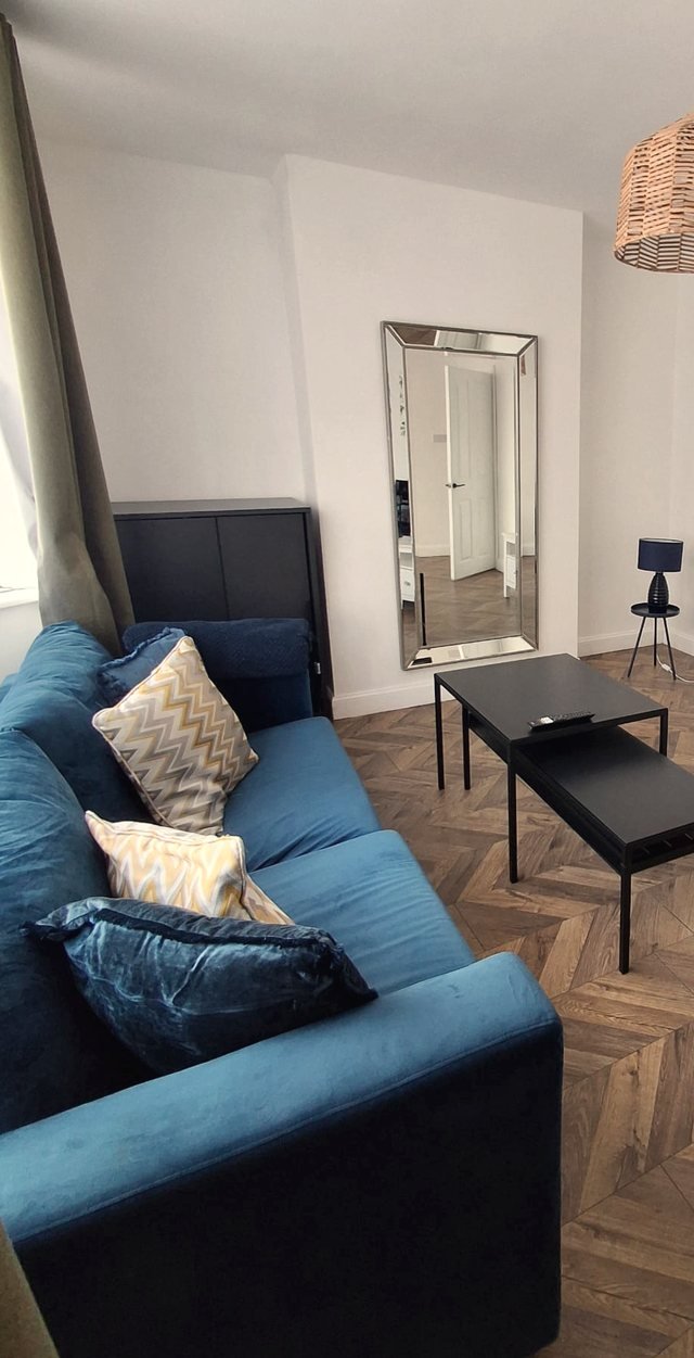 The sitting room at Oasis House: a blue velvet sofa with soft patterned cushions, a full-length mirror, a rattan pendant light and a herringbone wood floor.
