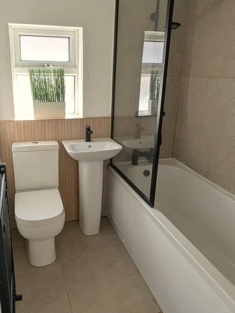 The family bathroom: a frosted window with a plant on the sill, wood-slat panelling, a basin and a bath with a black-framed screen.